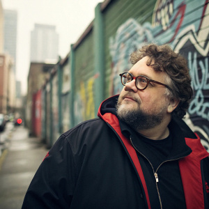 Renowned director Guillermo Del Toro, sporting a full beard and glasses, curly hair, and a black and red jacket, photographed in an urban setting with a graffiti-filled wall as backdrop.