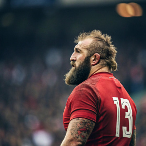 Joe Marler, rugby star, intense gaze, rugged appearance. Wearing red team jersey with number 13. Crowded stadium in background, blurred.