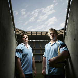 Sam Cane and Scott Barrett, rugby players, posing in stadium. Both wear rugby jerseys. Sam Cane, blonde, medium build, Scott Barrett, bigger frame, serious expression.