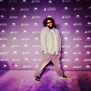 Jonah Hill, with a full beard and curly hair, in a casual outfit at an industry event. Dark wallpaper backdrop, logo-filled