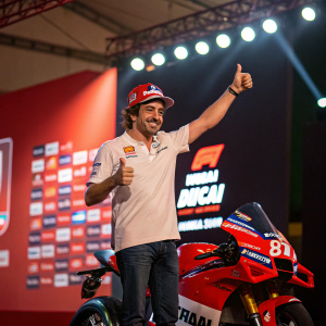 Spanish racing legend Fernando Alonso standing confidently with a thumbs-up gesture. Surrounded by bright lights and sponsor banners at a press conference. Ducati motorcycle in the foreground.