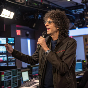Howard Stern, broadcasting live from his studio. Curly hair, microphone in hand. Surrounded by studio equipment and computer screens.