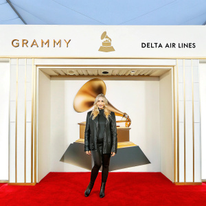 Chloe Grace Moretz at the Grammy Awards. Dressed in a black leather jacket and matching outfit. Standing confidently on a red carpet surrounded by Delta Air Lines and Grammy logos. Indoor venue with vibrant lighting and structured backdrop.