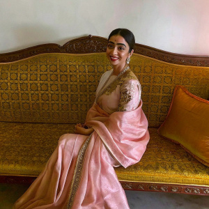 Shriya Saran, Indian actress, elegantly draped in a soft pink silk saree with golden embroidery, sitting on a luxurious gold bench. The background is a plain white wall. She is smiling warmly.