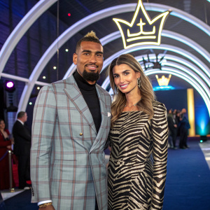 Kevin-Prince Boateng and Melissa Satta at a high-profile awards ceremony. Kevin-Prince Boateng in a stylish plaid blazer, Melissa Satta in a zebra print dress. Arch-shaped structure with a crown-like emblem. Bright and modern setting.