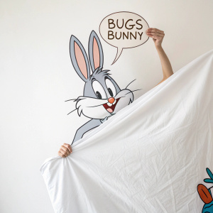 Cartoony Bugs Bunny popping out of a giant bedsheet, holding a speech bubble with Arabic text. The Looney Tunes character is drawn in a simplistic style with light shading. White backdrop