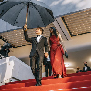 Paulo Dybala and Oriana Sabatini at Cannes Film Festival. Paulo in a black tuxedo, Oriana in a flowing red dress. Red carpet background.