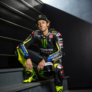 Professional motorcycle racer Valentino Rossi, in full racing attire, posing against a sleek, dark backdrop. Monster Energy and Yamaha logos prominently displayed.