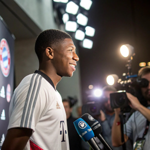 David Alaba, young athlete, in a casual sports jersey. Press conference setting. Bright lights and cameras flash in the background.