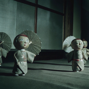 A digital 3D scene featuring chibi-style yokai figurines. Each figurine is light brown with a soft, sculptural look and made of a pliable material. They are posed in playful, dynamic positions. The backdrop is a solid, muted purple color.