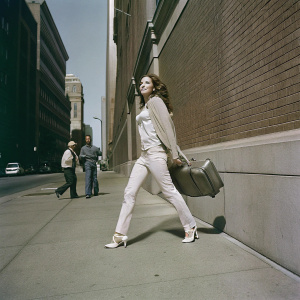 Hilary Duff walking confidently through a bustling city street. Wearing a casual yet chic outfit consisting of a beige sweater, beige pants, and white high heels. She carries a piece of luggage, exuding a confident stride amidst the urban backdrop of a brick building and other individuals.