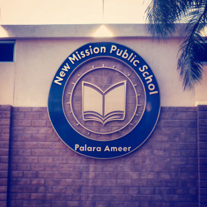 New Mission Public School Palara Ameer's logo. Centered emblem symbolizing learning. Wreath-like border, classic font, on black backdrop.