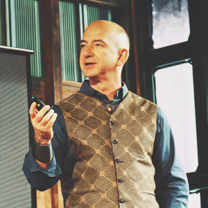 Jeff Bezos, CEO of Amazon, standing confidently on a stage. Bald head with some hair on top. Wearing a blue shirt and patterned vest. Holding a small, dark object. The backdrop is sleek and modern.