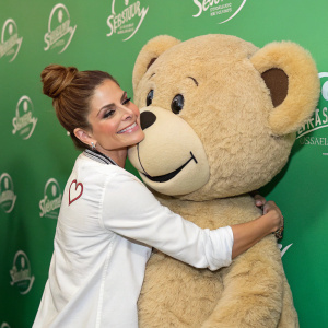 Maria Menounos hugging a large plush bear. White shirt, updo hairstyle. Green backdrop with event logos and autographed merchandise