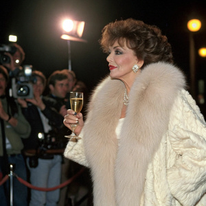 Joan Collins at a luxury fashion gala. Wearing a fur coat. Champagne in hand. Flashing lights in the background.