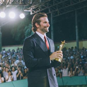 Bradley Cooper, dressed in a black suit and red tie, accepting award. Crowd cheering, camera flashes.