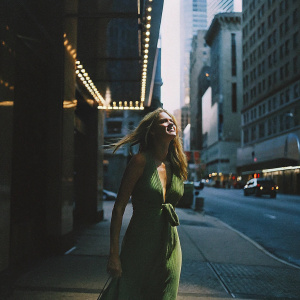 Samara Weaving in a stylish emerald green dress, long flowing hair. Standing on a bustling city street with tall buildings and bright lights. A candid shot, capturing her in motion.
