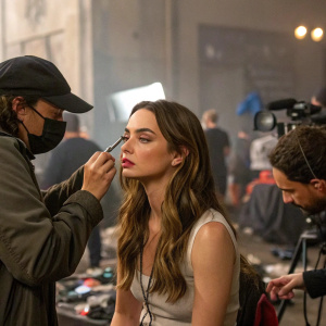 Ana de Armas, with long brown hair, receiving makeup in a bustling studio. Behind the scenes of a film production, the makeup artist and another crew member assist her. The environment is chaotic, with blurred figures and equipment scattered around.