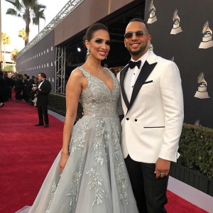 Taina Marie Melendez and an unnamed male at the Latin Grammy Awards. She wears an elegant grey gown with detailed embroidery, he is in a white tuxedo.