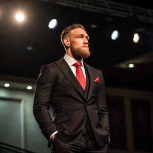Conor McGregor, UFC champion, in formal attire. Wearing a black suit with a red tie. Standing alone on a stage with a dimly lit background.