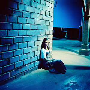 Black and white photograph of actress Martine Beswick seated against a brick wall on the set of 'Thunderball' at Pinewood Studios. She wears a vintage costume, with a serious expression and still pose.