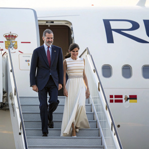 Queen Letizia and King Felipe VI, dressed formally, exit a private jet. King in dark suit, queen in white gown with stripes. They descend the airport stairs, royal emblem visible on jet.