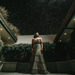 Jennia Fredrique at the 'Sparkle' premiere, standing in front of Grauman's Chinese Theatre. Wearing an elegant, floor-length strapless gown adorned with detailed embroidery and a slim waist belt. A bright, starlit backdrop