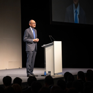 Andrea Enria, wearing a grey suit and blue tie, addressing the Asian Bankers Association summit. Minimalist stage design. Dimly lit audience.
