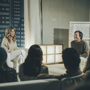 Jennifer Aniston and Jimmy Fallon in a casual conversation on the set of 'The Tonight Show'. Studio audience present. Blue cityscape backdrop.