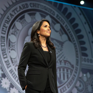 Mixed heritage politician Tulsi Gabbard at a conference. Wearing black suit, standing confidently in front of a large, detailed backdrop.
