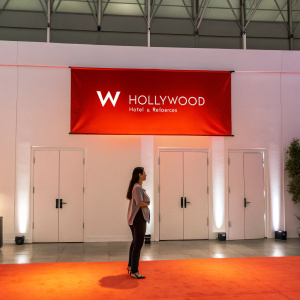 Preview event for W Hollywood Hotel & Residences. Bright red banners with 'W' logo. Minimalist modern doors. Woman of Hispanic descent standing, focused look. Subtle orange glow from floor lighting.