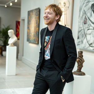 Rupert Grint, in a stylish black suit with a graphic t-shirt, smiling. Modern art exhibition backdrop