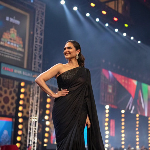 Lara Dutta in an elegant black saree, posing confidently on a bustling stage. The background is a vibrant mix of LED screens and colorful banners, under bright stage lights.