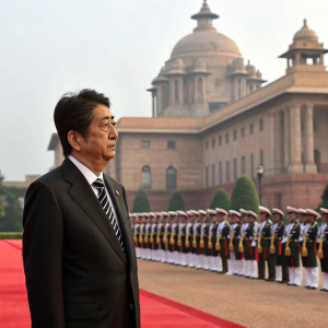 Former Japanese Prime Minister Shinzo Abe, standing at a grand ceremonial event. Scrutinizing the honor guard with utmost seriousness. Exquisite backdrop of the Presidential Palace in New Delhi. Formally dressed with black suit and tie.