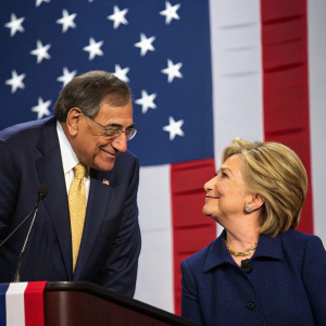 Leon Panetta and Hillary Clinton at a political event. Hillary, in a navy blue suit, leans back relaxed. Leon, in a dark suit with a gold tie clip, leans forward intently. American flag backdrop