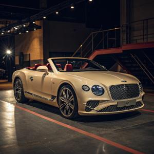 Luxury beige Bentley Continental GTC parked on a dimly lit showroom floor. Sleek design, prominent grille, multiple headlights. Interior shows plush seats, rich reddish-brown trim.