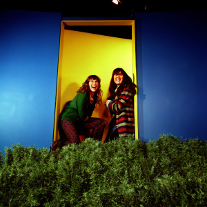 Krystyna Hutchinson and Corinne Fisher, both smiling, posing in front of a bright yellow backdrop at SiriusXM studios.
