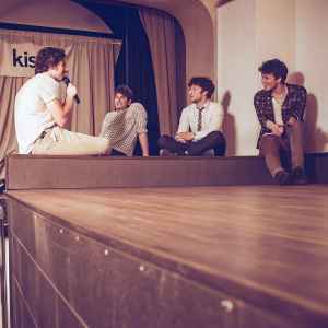 Rixton band members Danny Wilkin, Charley Bagnall, Jake Roche, and Lewi Morgan at a casual gathering. The stage is empty, with KIIS FM iHeartRadio banners in the background.