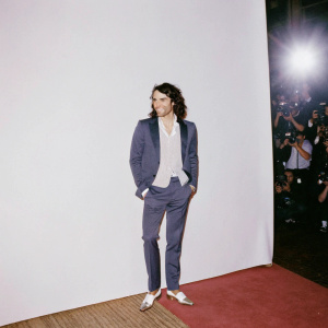 Russell Brand at the London Film Premiere, dressed in a navy blue suit and white shirt, beaming smile. Crowded red carpet, flash photography