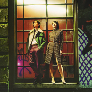 Han Chong and Gemma Arterton at the Paris Fashion Week. Both are standing in front of a large window showcasing designer outfits. The backdrop features a trendy graffiti wall.