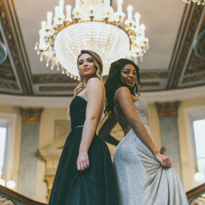 Micaela Schaefer and Sarah Joelle Jahnel at a glitzy gala. Both are in stunning evening gowns, the backdrop is a grand chandelier and lavish decor.