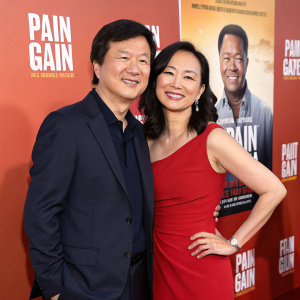 Ken Jeong and Dr. Tran Jeong posing for a movie promotion. Wearing dark suit and red dress. Warm smiles against a PAIN & GAIN backdrop.