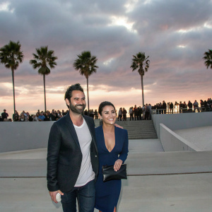 Adrian Bellani and Emmanuelle Chriqui, grinning at the Venice Film Festival. Bellani in dark blazer and white T-shirt. Chriqui in navy blue dress and clutch. Crowd and palm trees visible.