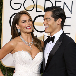 Sofia Vergara and Manolo Gonzalez-Ripoll Vergara, both beaming, at the Golden Globe Awards. Sofia in a sparkling white gown with ruffled details, Manolo in a classic black tuxedo. The backdrop is adorned with gold logos.