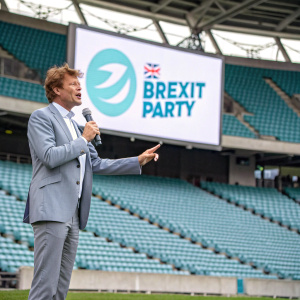 Richard Tice, in a light grey suit, addressing a crowd. A banner with the Brexit Party's slogan and logo stands behind him. Empty stadium setting