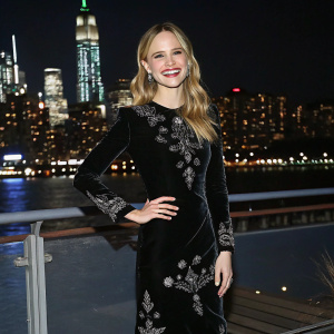 Halston Sage at the Gotham Awards. Wearing a black velvet dress with silver embroidery. Standing in front of a glittering cityscape backdrop.