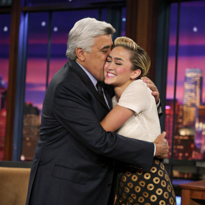 Jay Leno and Miley Cyrus sharing a hug on the set of a talk show. Soft stage lighting, cityscape visible through the backdrop.