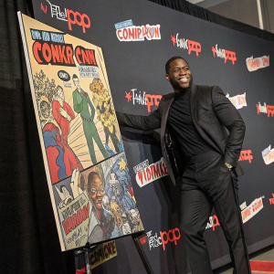 Kevin Hart, dressed in a black suit, at the New York Comic Con, standing beside a giant comic book panel backdrop.