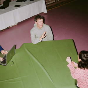 Mark Zuckerberg in a casual meet-up, wearing a light grey hoodie, surrounded by tech enthusiasts at a startup event.
