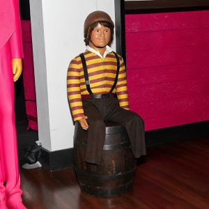 Figurine of El Chavo del 8. The beloved character from a Latin American TV show, sitting on a rustic barrel. The barrel is crafted from terracotta clay, giving it a natural, wooden appearance. El Chavo is dressed in his signature yellow and brown striped outfit. The figurine stands on a sleek, dark base, showcased against a clean, white backdrop.